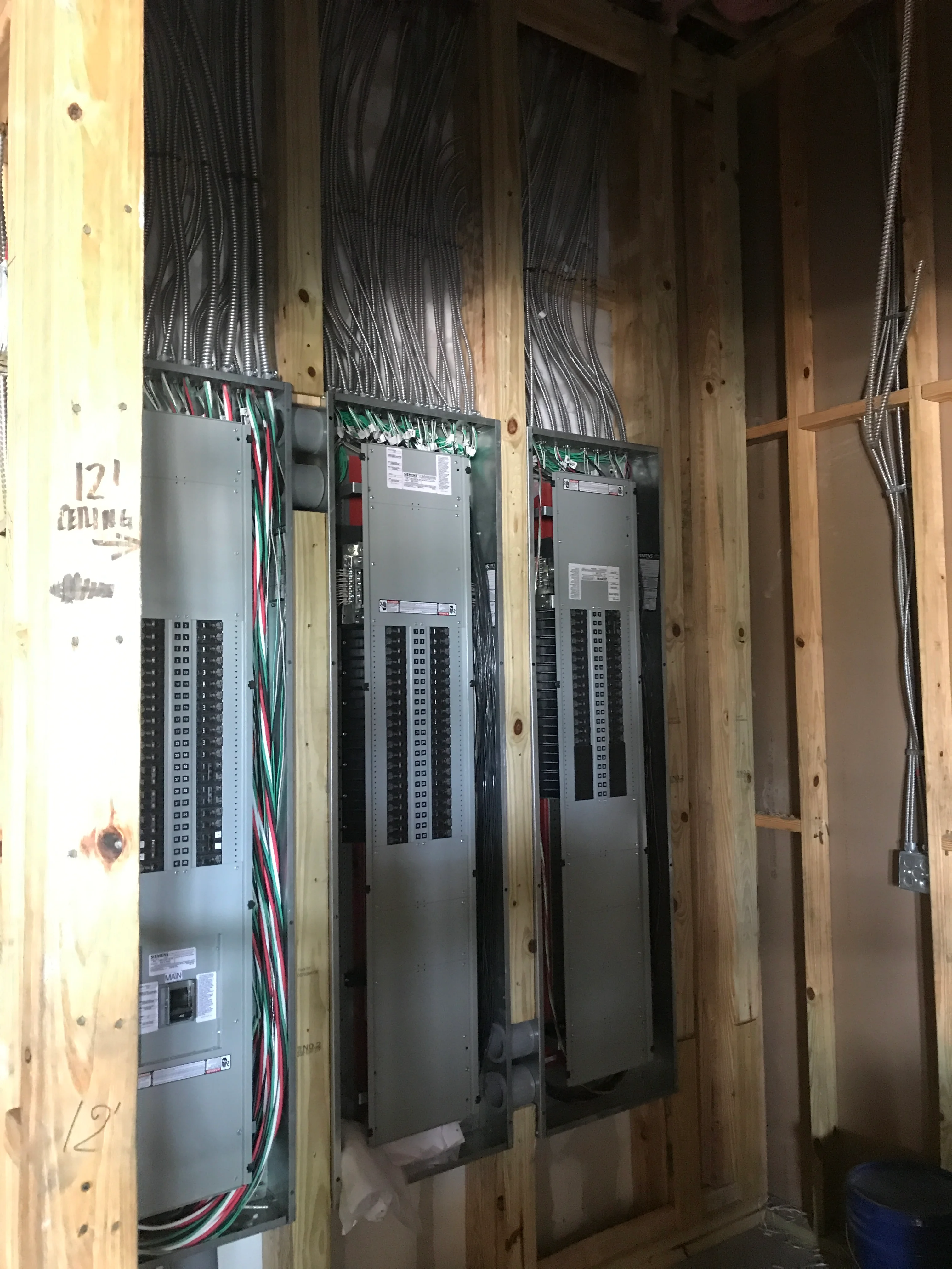 Three electrical distribution panels with organized metallic conduit and color-coded wiring in framed wall cavity