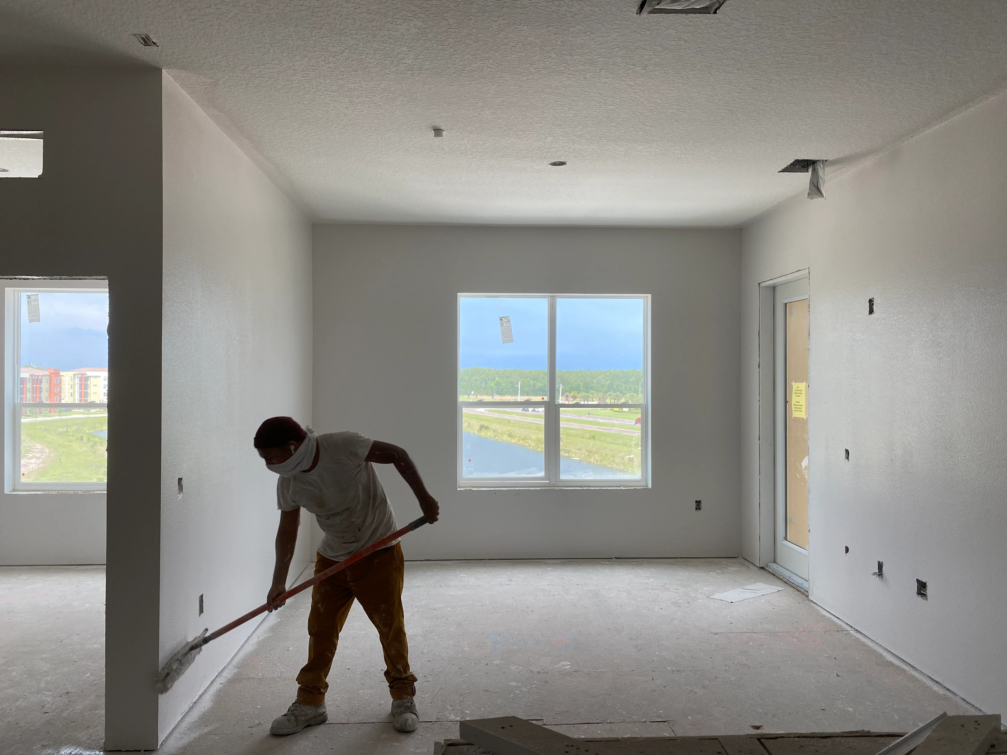 Worker cleaning concrete floor in newly finished drywall interior with large windows