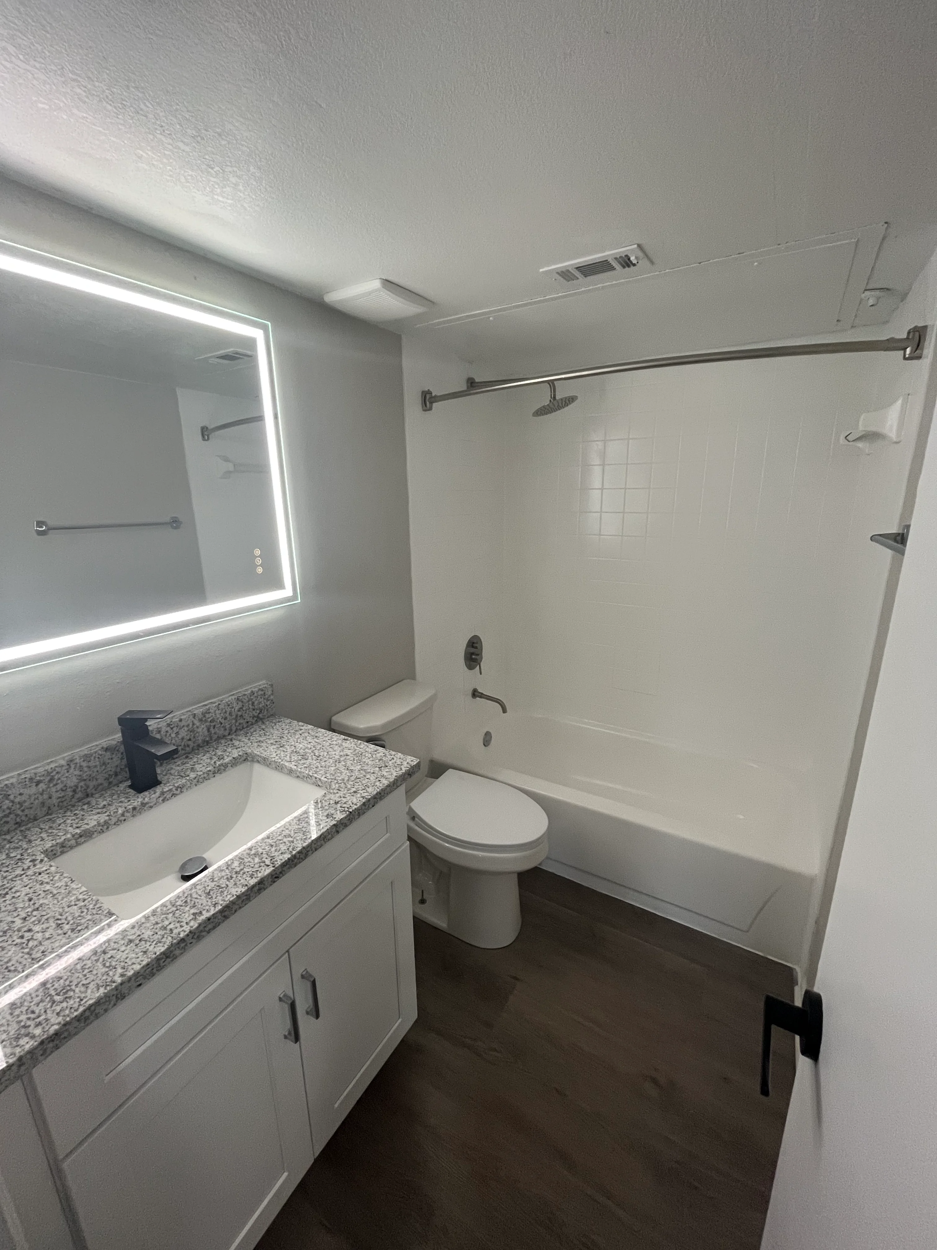 Modern bathroom with granite vanity, LED mirror, white shower surround, and dark wood flooring